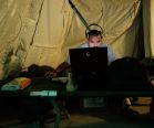 Illustration: A private civilian contractor works with his laptop inside a transit tent.